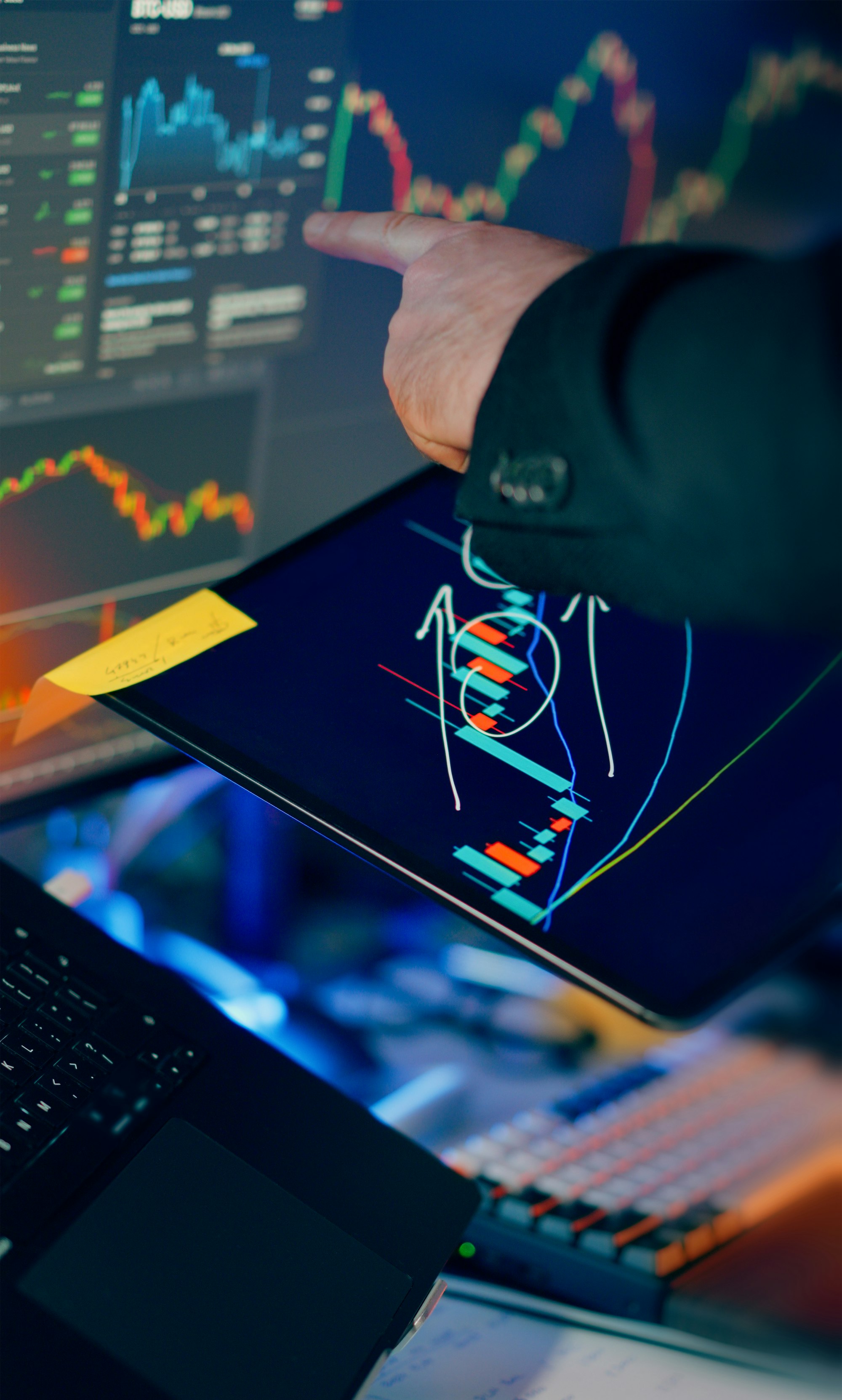 A trader's hand pointing at candlestick charts across several screens